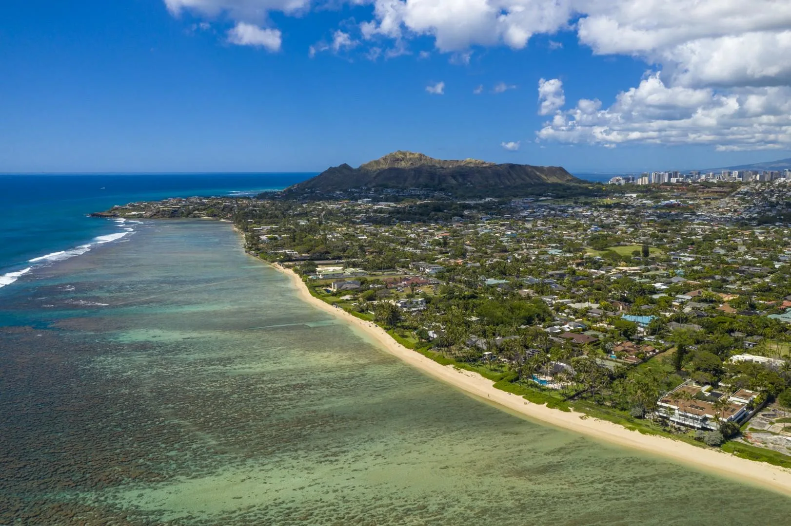 Aerial coastal view of a sandy shoreline, reef, and town beneath a mountain.