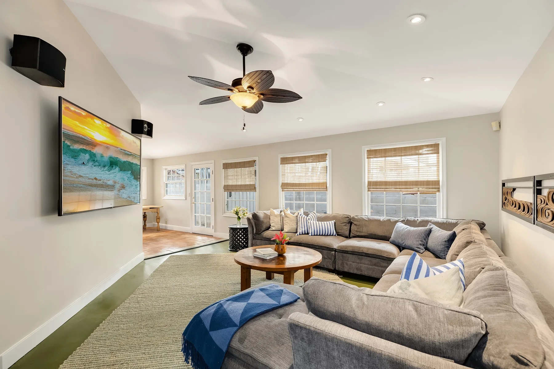 A bright living room with sectional seating, a coffee table, and windows with blinds.
