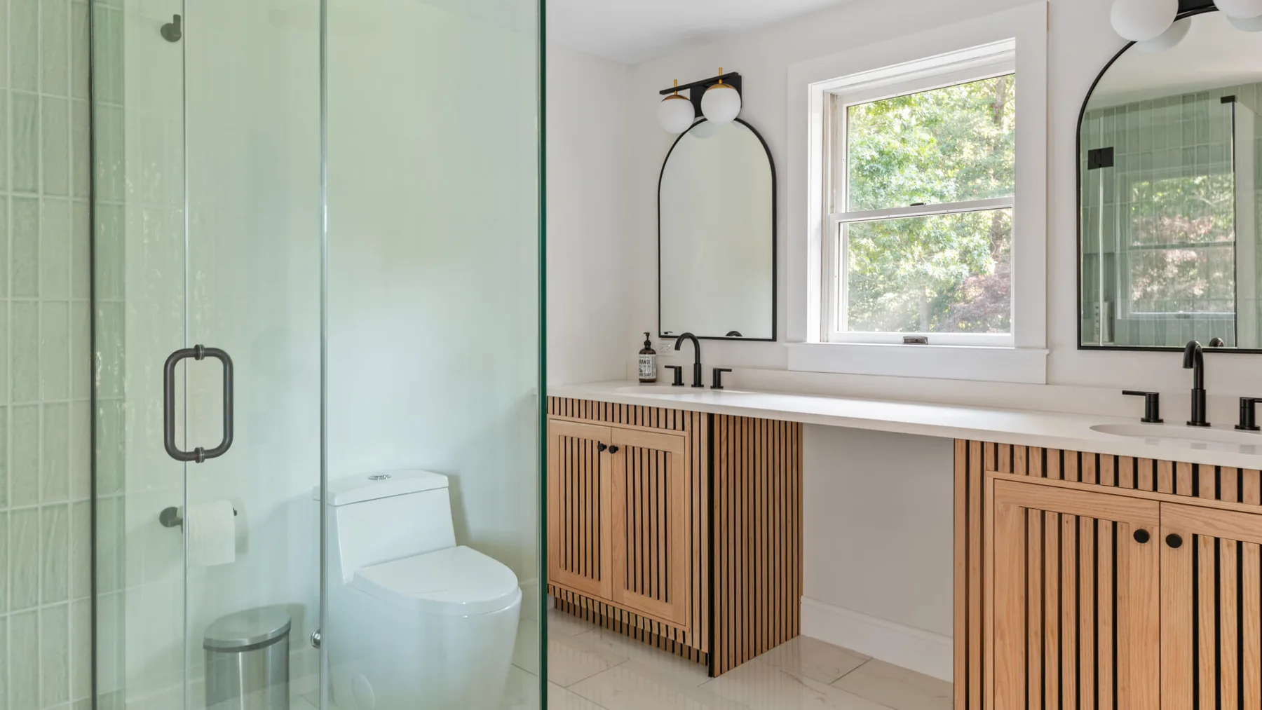 A bright bathroom has a glass shower, a toilet, and a double vanity beneath a window.