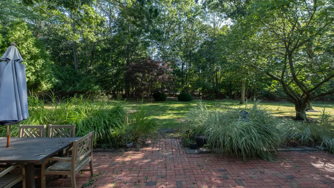 A brick patio opens onto a leafy garden with trees and ornamental grasses.