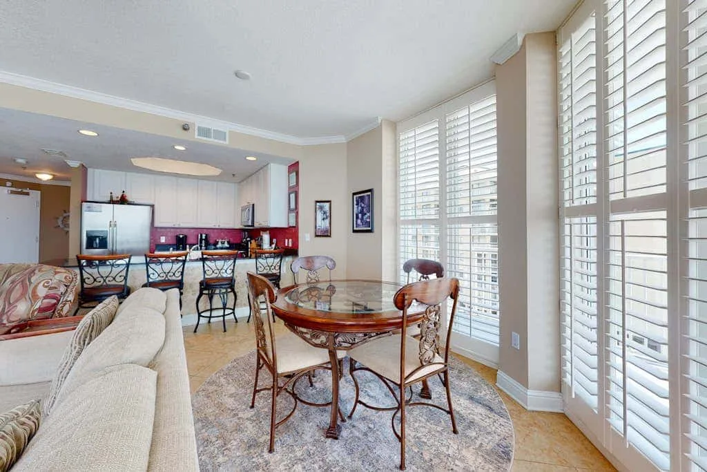 A bright living and dining area with a round table beside large shuttered windows.
