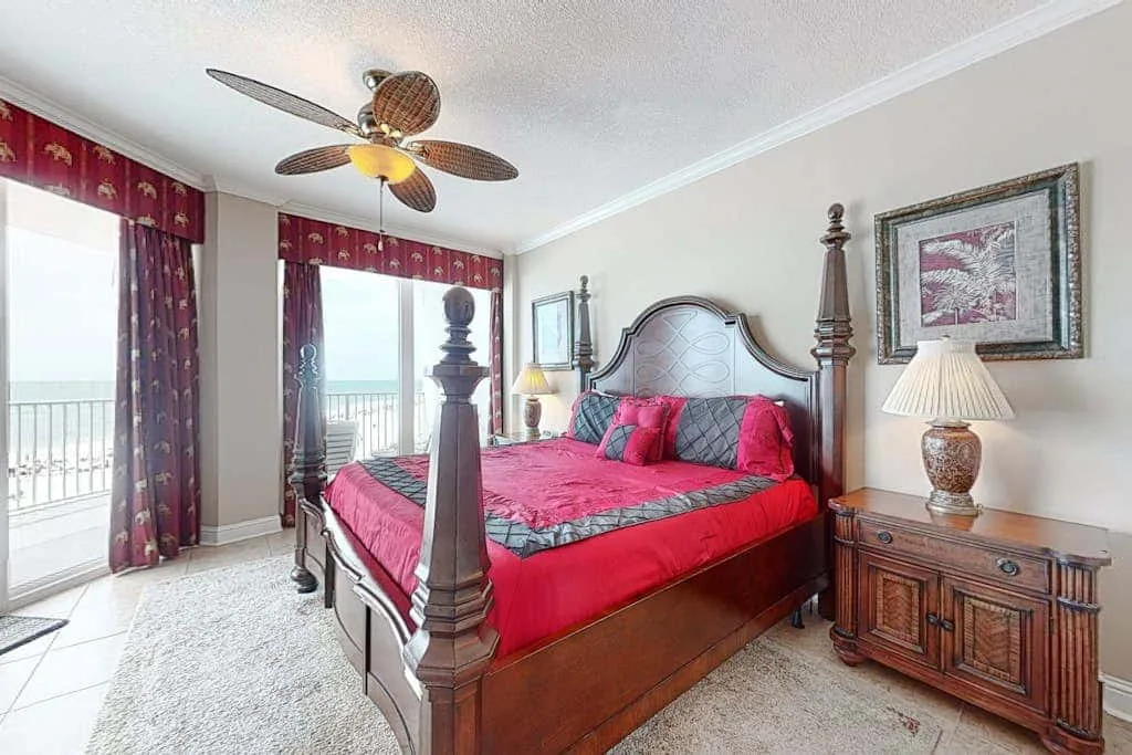 A bedroom with a four-poster bed dressed in red bedding beside bright windows.