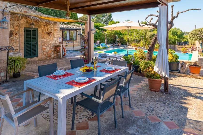 Una zona de comedor al aire libre con sombra da a una piscina y un jardín.