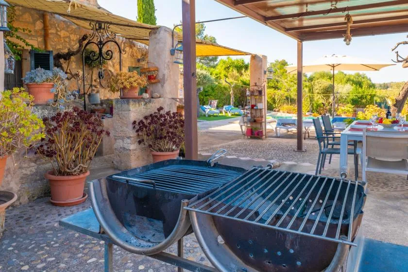 Un patio exterior con dos parrillas en primer plano y asientos bajo una zona techada.