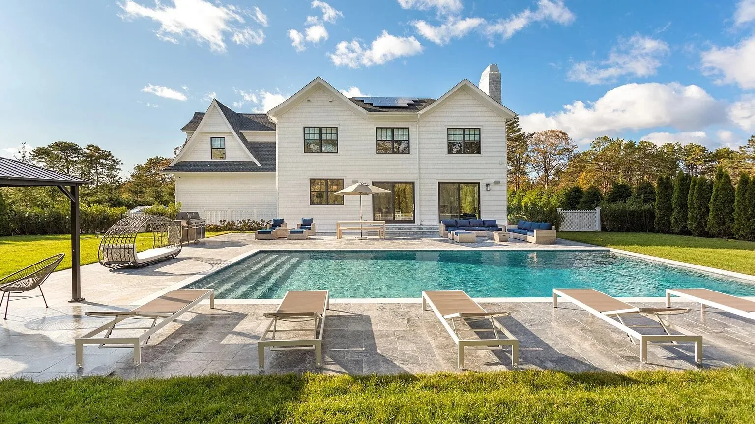 A large house stands behind a swimming pool and patio seating.