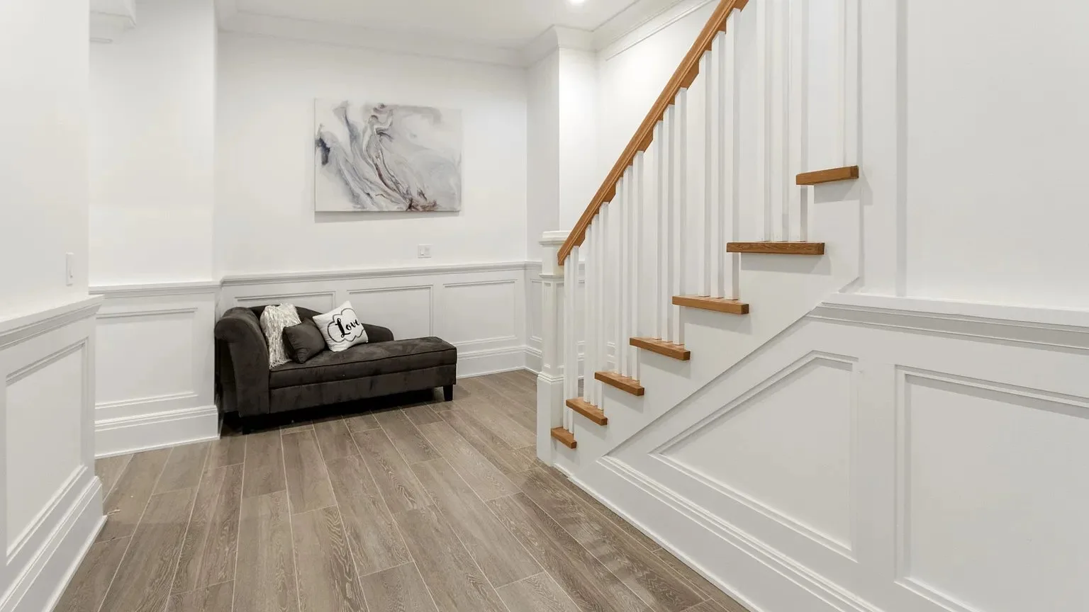 Una luminosa zona de estar con un sofá oscuro bajo un cuadro abstracto, junto a una escalera.