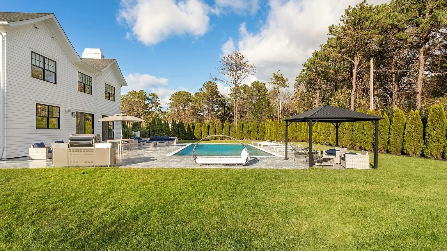A lawn and swimming pool beside a white house, with a shaded seating area.