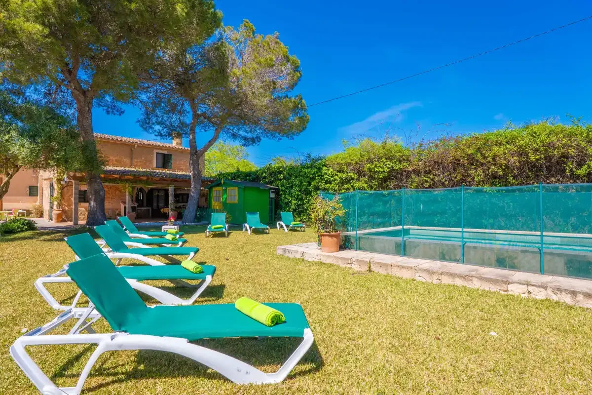 A sunny outdoor pool area with green loungers beside a fenced swimming pool.