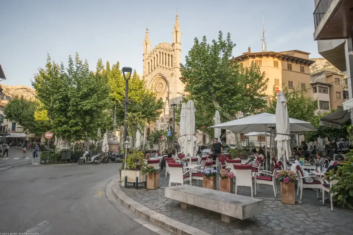 A town square with outdoor café seating and a tall stone church behind the trees.