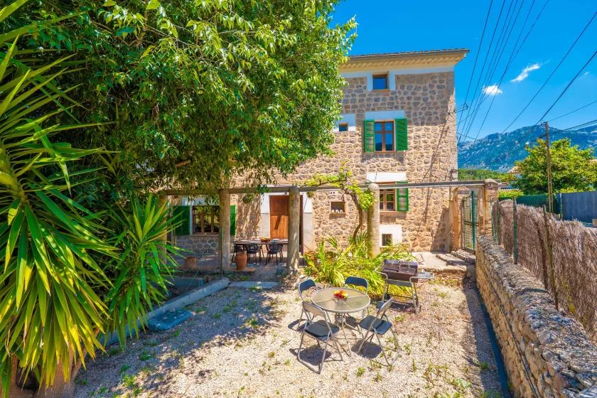 A stone house overlooks a gravel patio with outdoor seating and leafy plants.