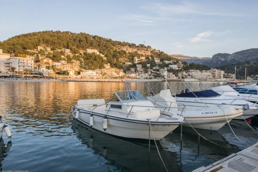Small boats are moored along a waterfront, with hillside buildings across the water.