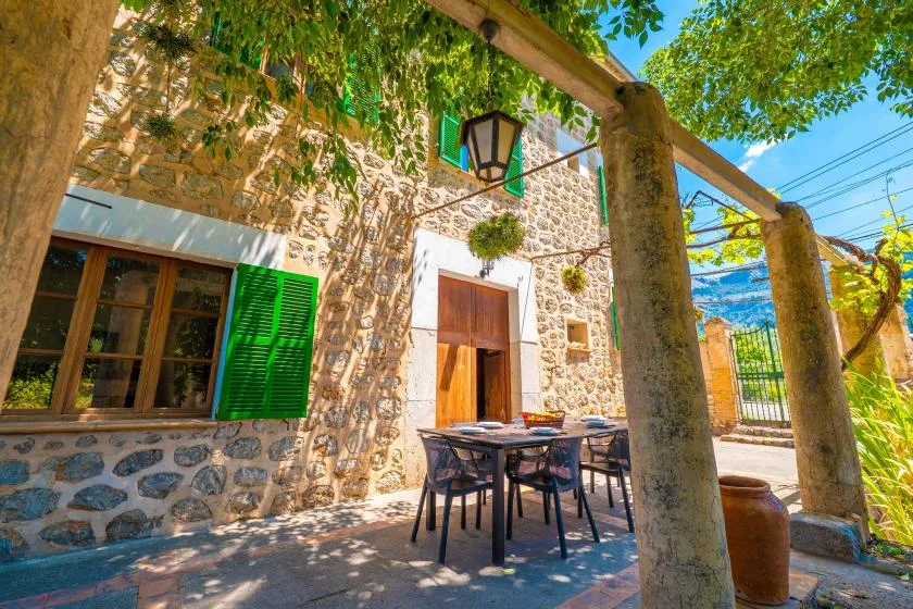 A stone house patio with outdoor seating beneath a leafy wooden pergola.