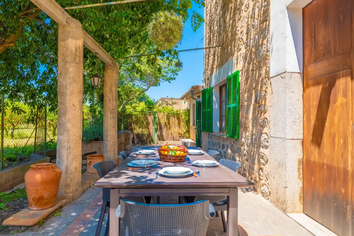 An outdoor dining table sits beside a stone building under leafy trees.
