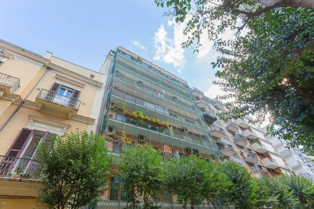 A city apartment building with balconies and leafy trees in the foreground.