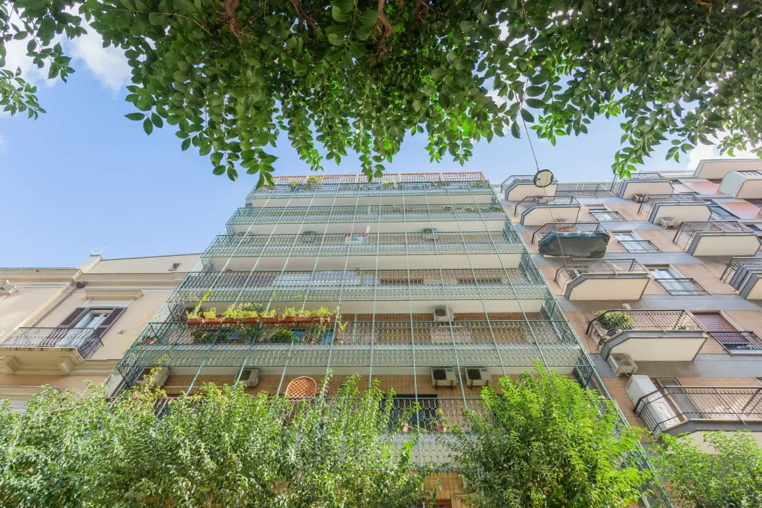 A multi-story apartment building is viewed from below through leafy tree branches.