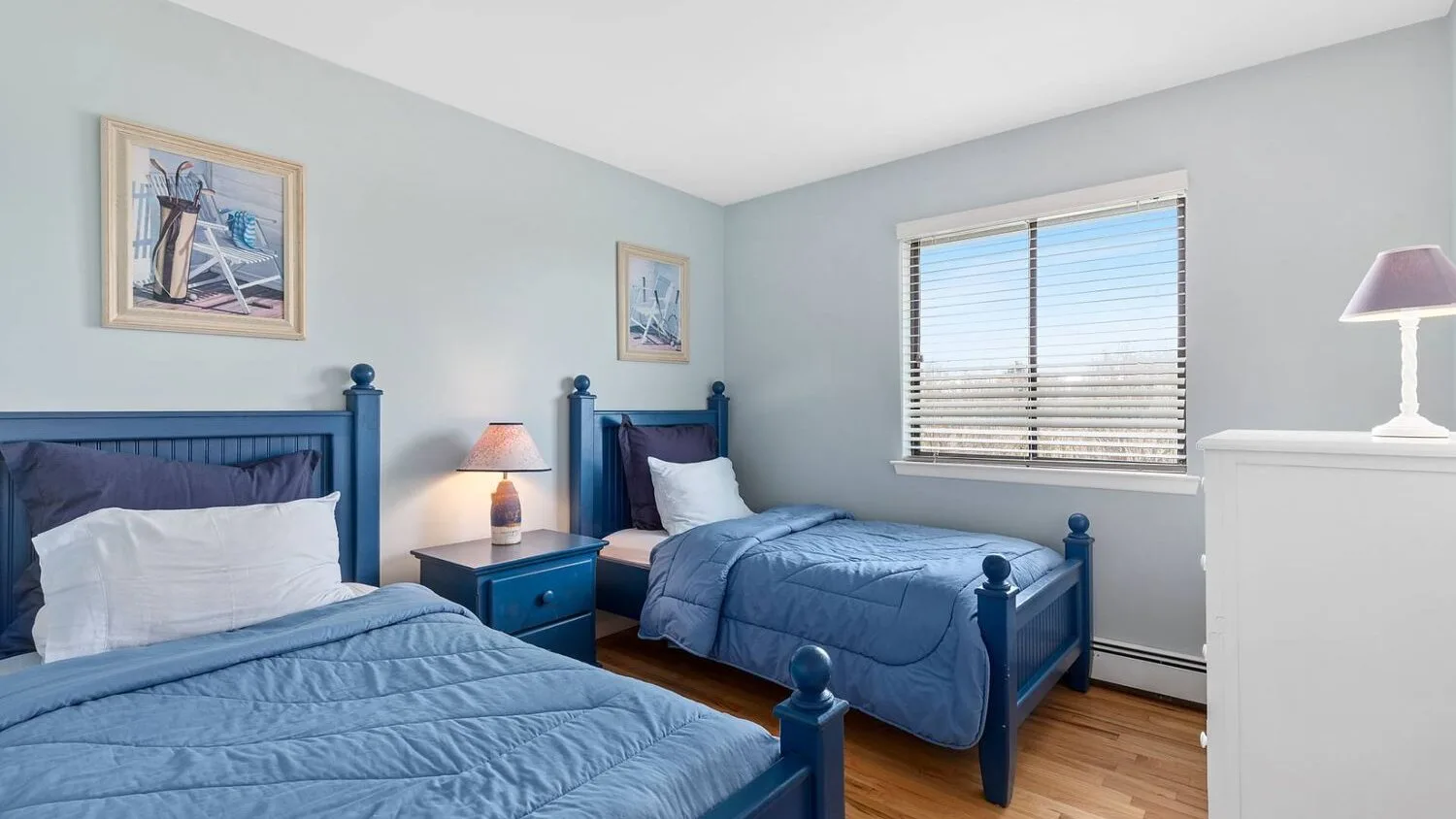 A bedroom with two blue beds, a shared nightstand, and a window with blinds.