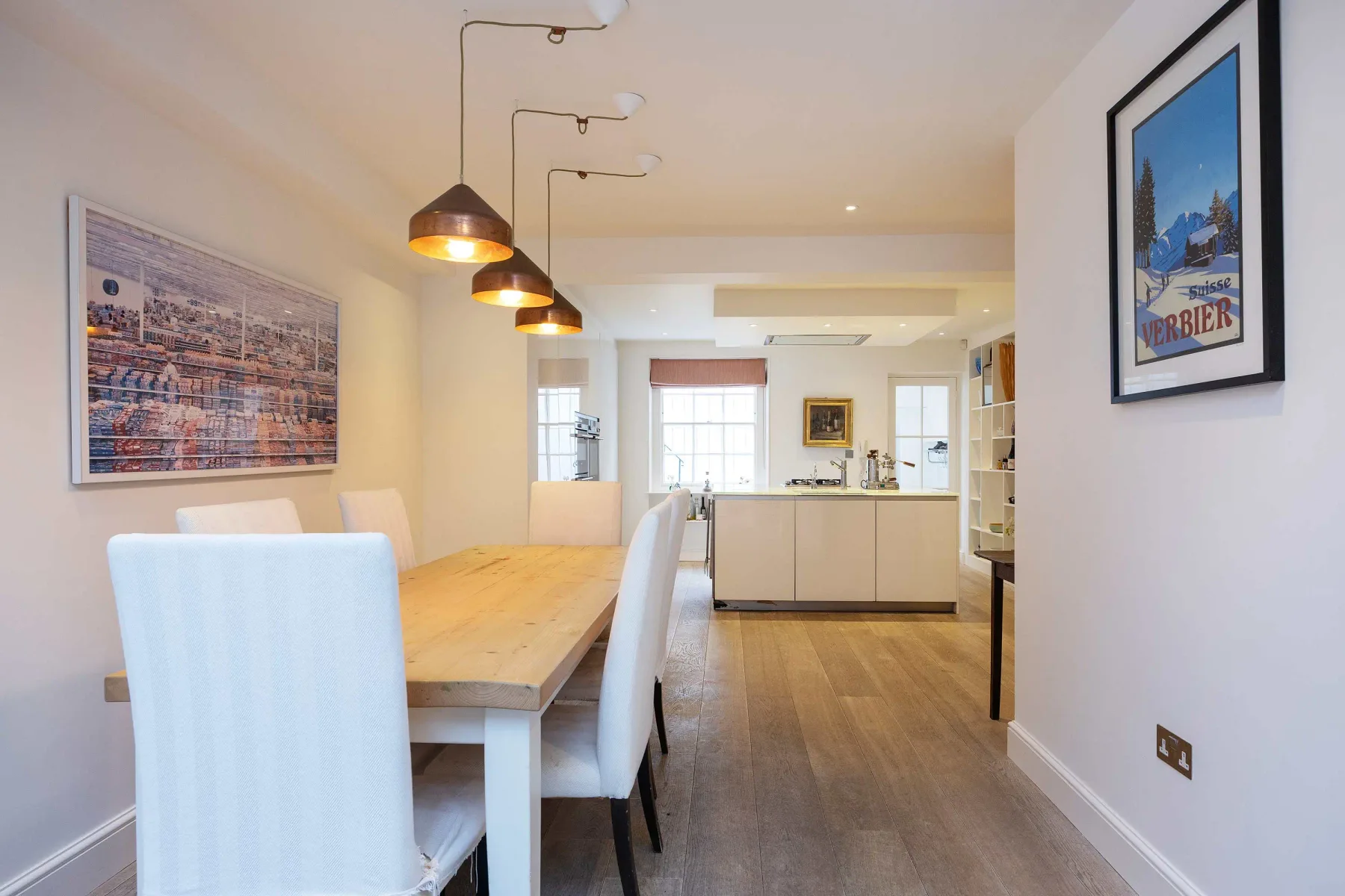 Un comedor con una mesa larga y lámparas colgantes se abre a una cocina.