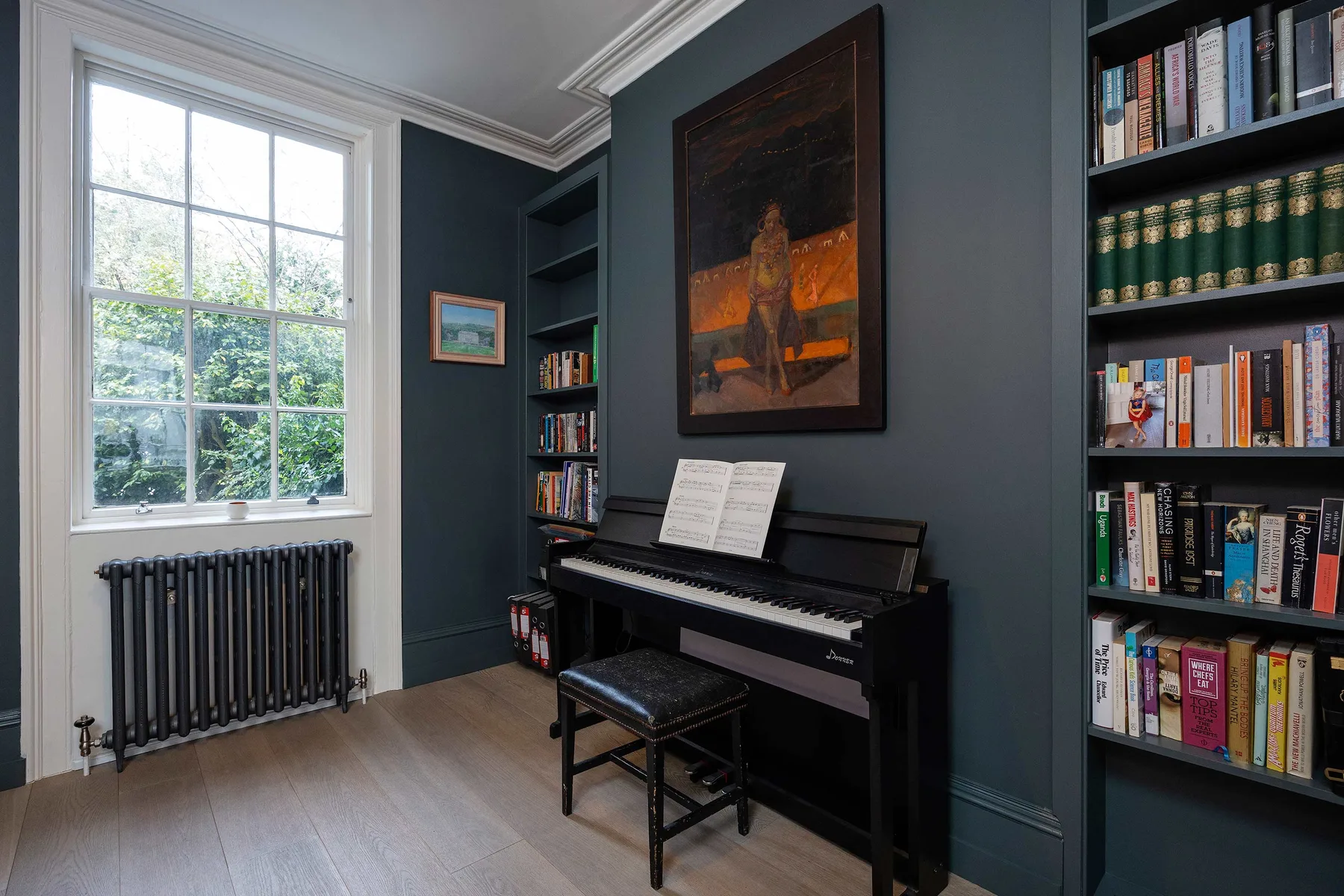 A dark-walled room with a piano, bookshelves, and a large window.