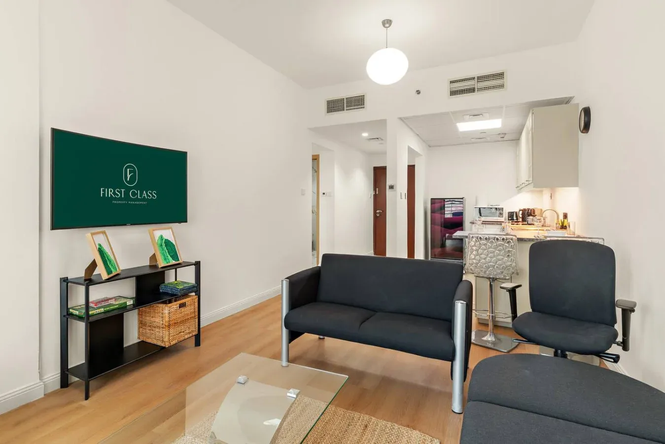 Una sala moderna con asientos oscuros, una mesa de centro de vidrio y una cocina compacta.