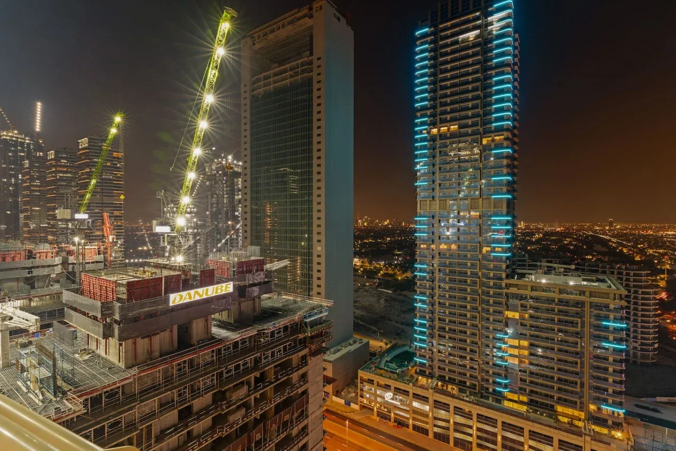 Panorámica nocturna de la ciudad con edificios altos iluminados y grúas de construcción.