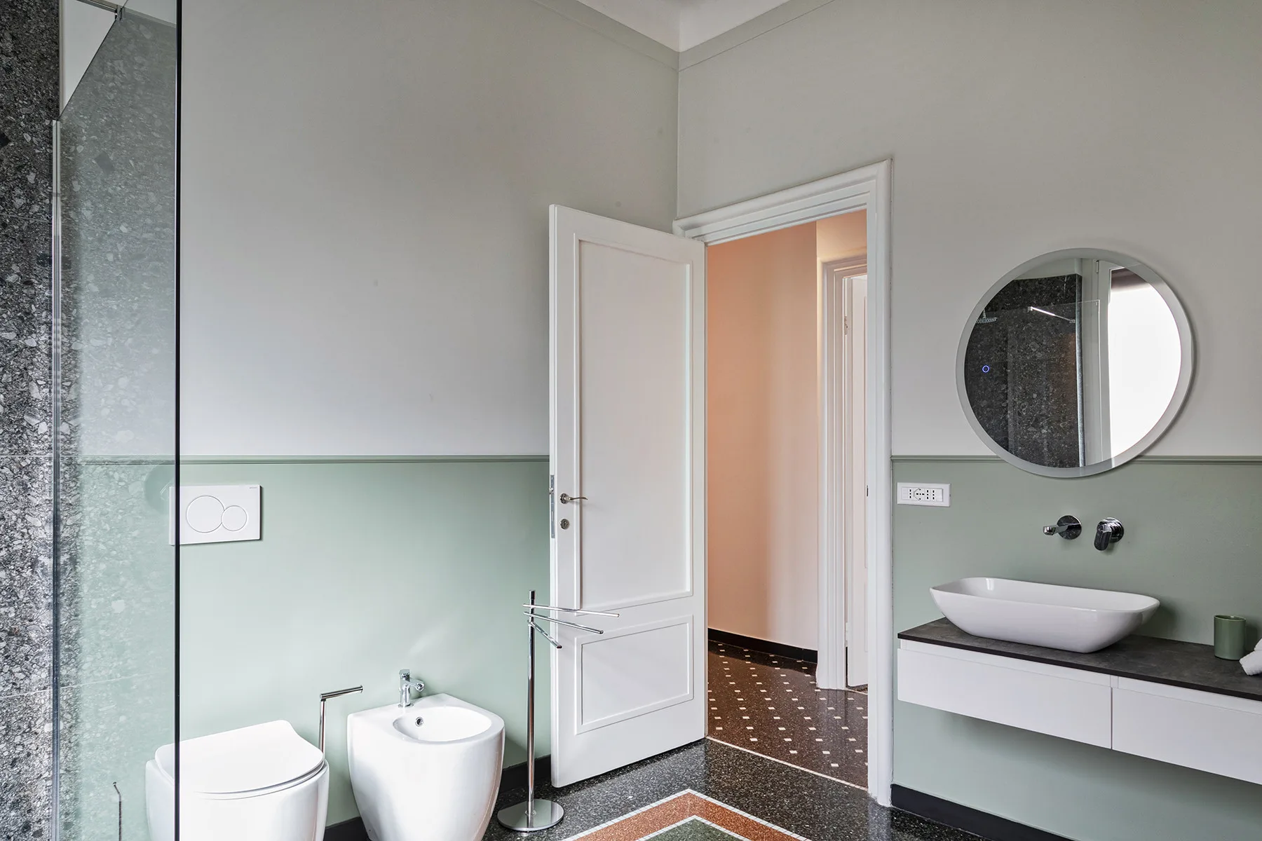 A bathroom with a glass shower, toilet, bidet, and sink beneath a round mirror.