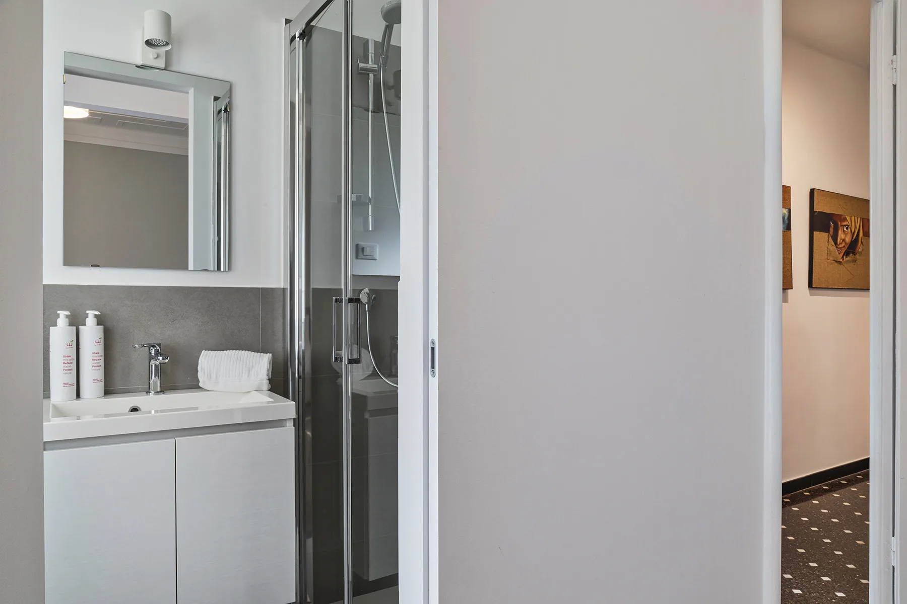 A bathroom with a sink, mirror, and glass shower beside an open doorway.