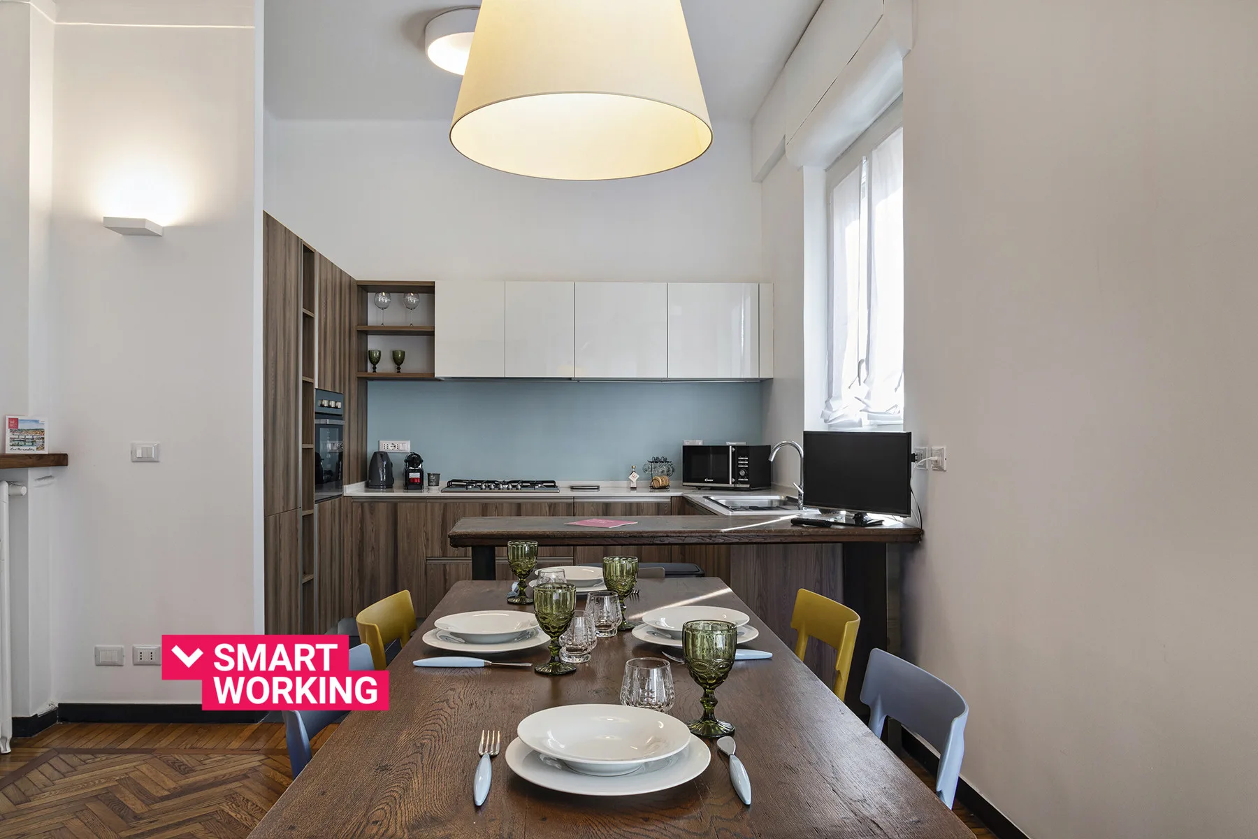 A dining table sits beside a modern kitchen with white cabinets and a large window.