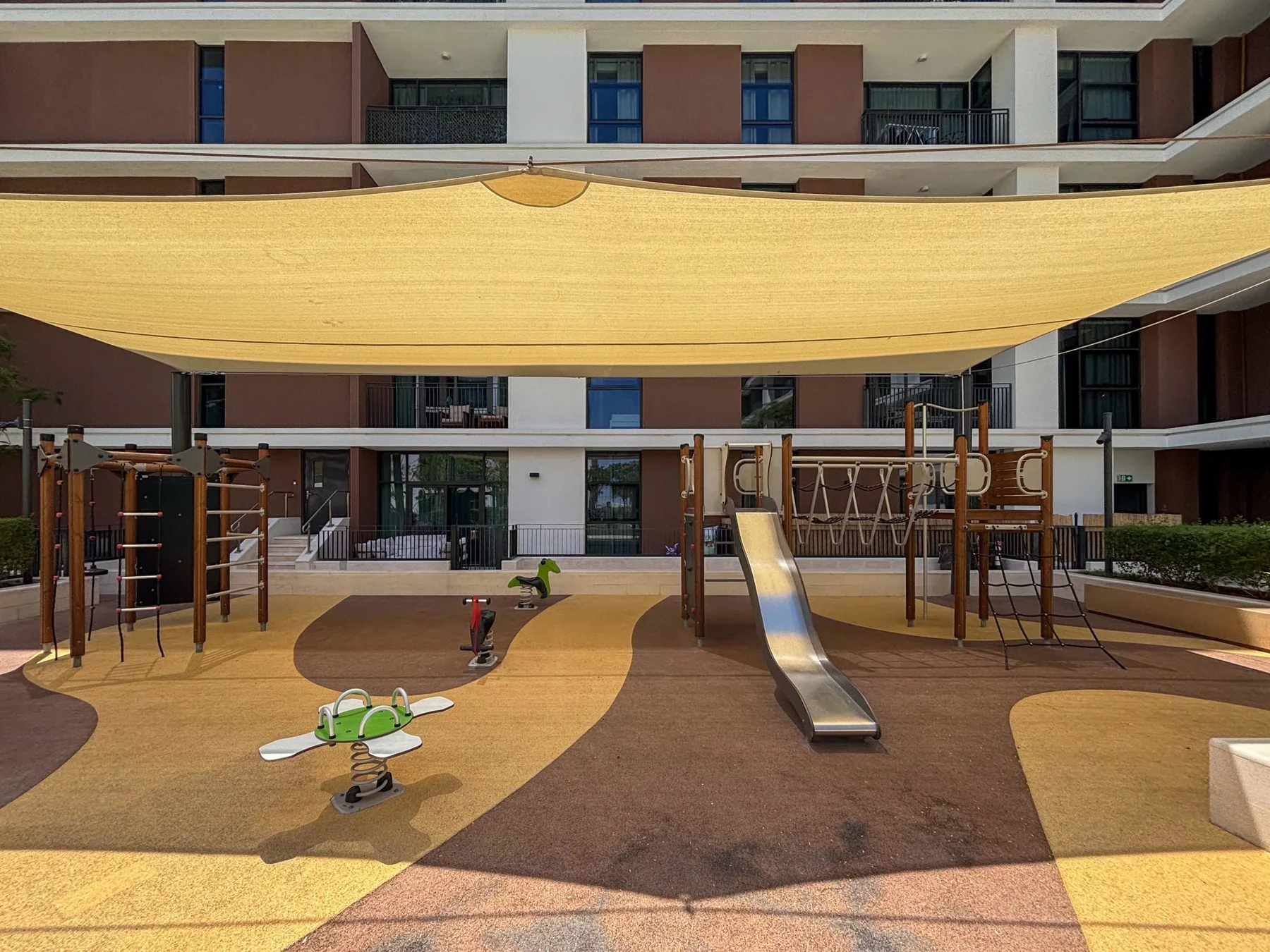 Un parque infantil sombreado con un tobogán y juegos para trepar frente a un edificio de apartamentos.