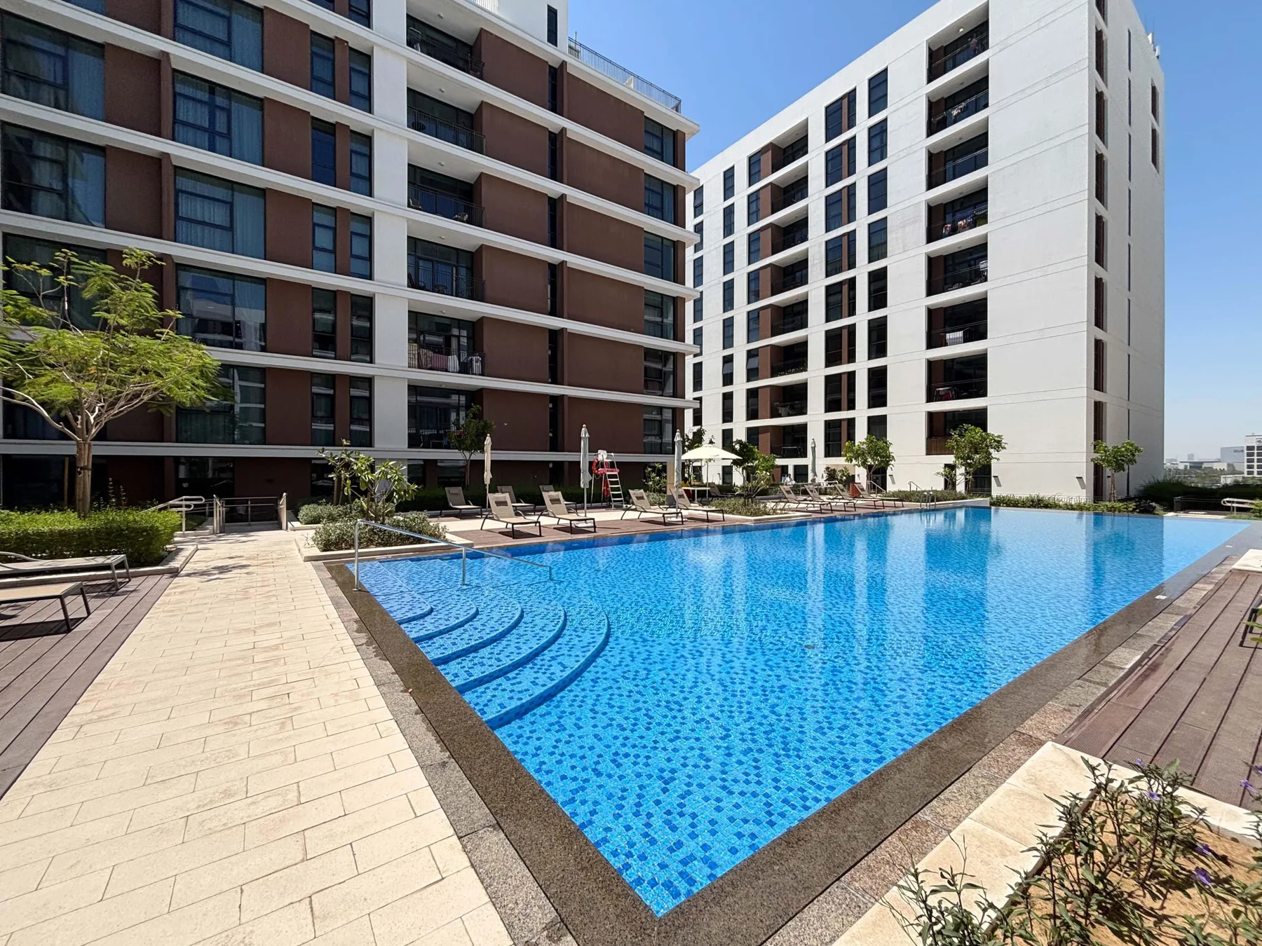 Una piscina se encuentra entre edificios de apartamentos modernos, con senderos pavimentados y asientos cerca.