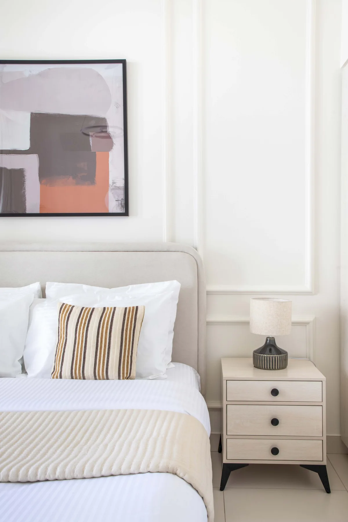 Un dormitorio con ropa de cama blanca, un cojín a rayas y una obra enmarcada.