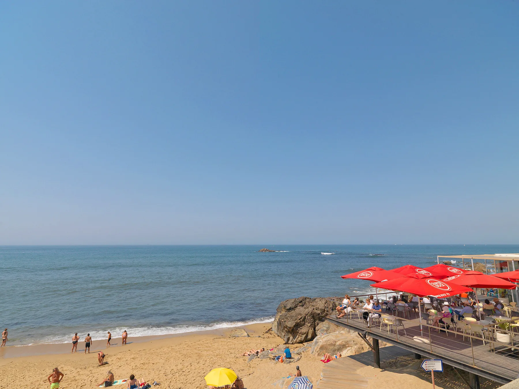 A sandy beach meets the sea beside a waterfront café terrace with red umbrellas.