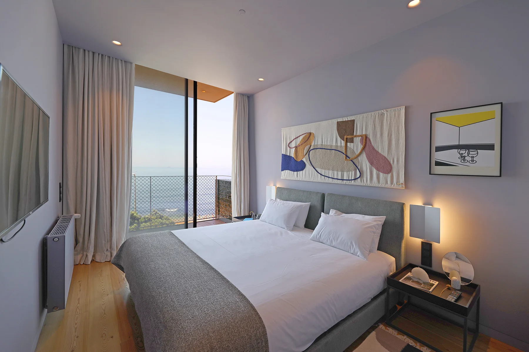 A bright bedroom with a bed beside a floor-to-ceiling window overlooking the sea.