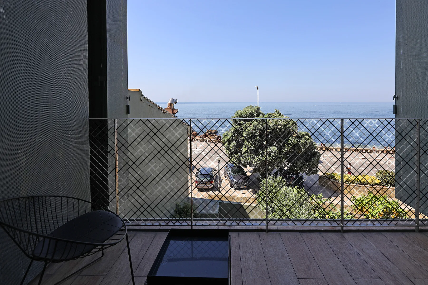 A balcony with outdoor seating overlooks the sea beyond a fenced area.