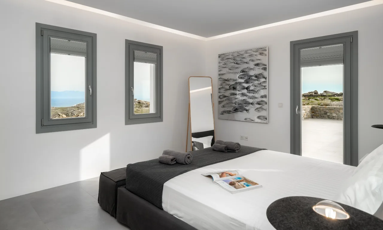 A bright bedroom with a low bed, dark bedding, and windows overlooking the landscape.