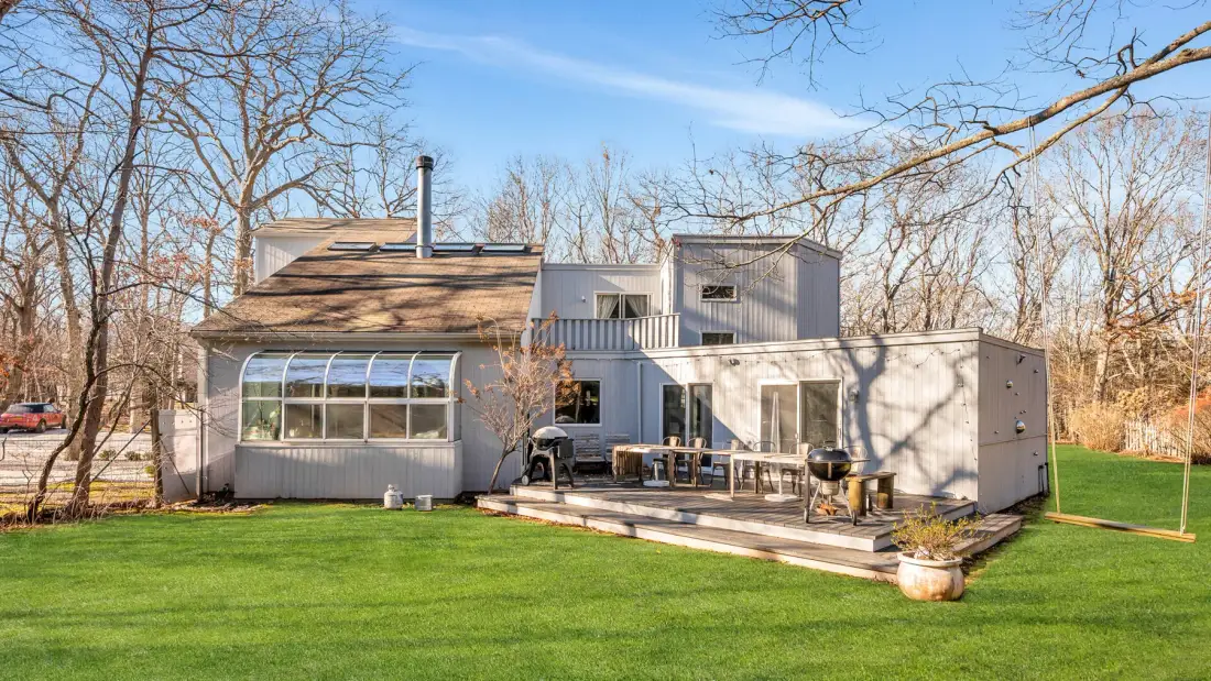 Una casa moderna con una terraza elevada da a un césped verde y árboles sin hojas.