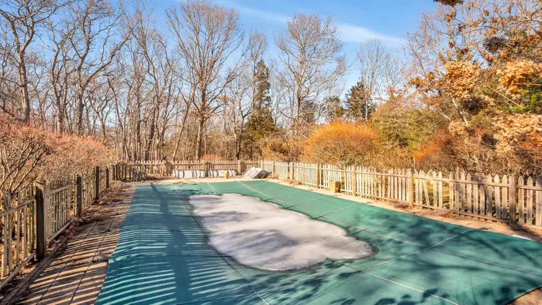 Una piscina exterior cercada, cubierta con una lona verde y rodeada de árboles otoñales.