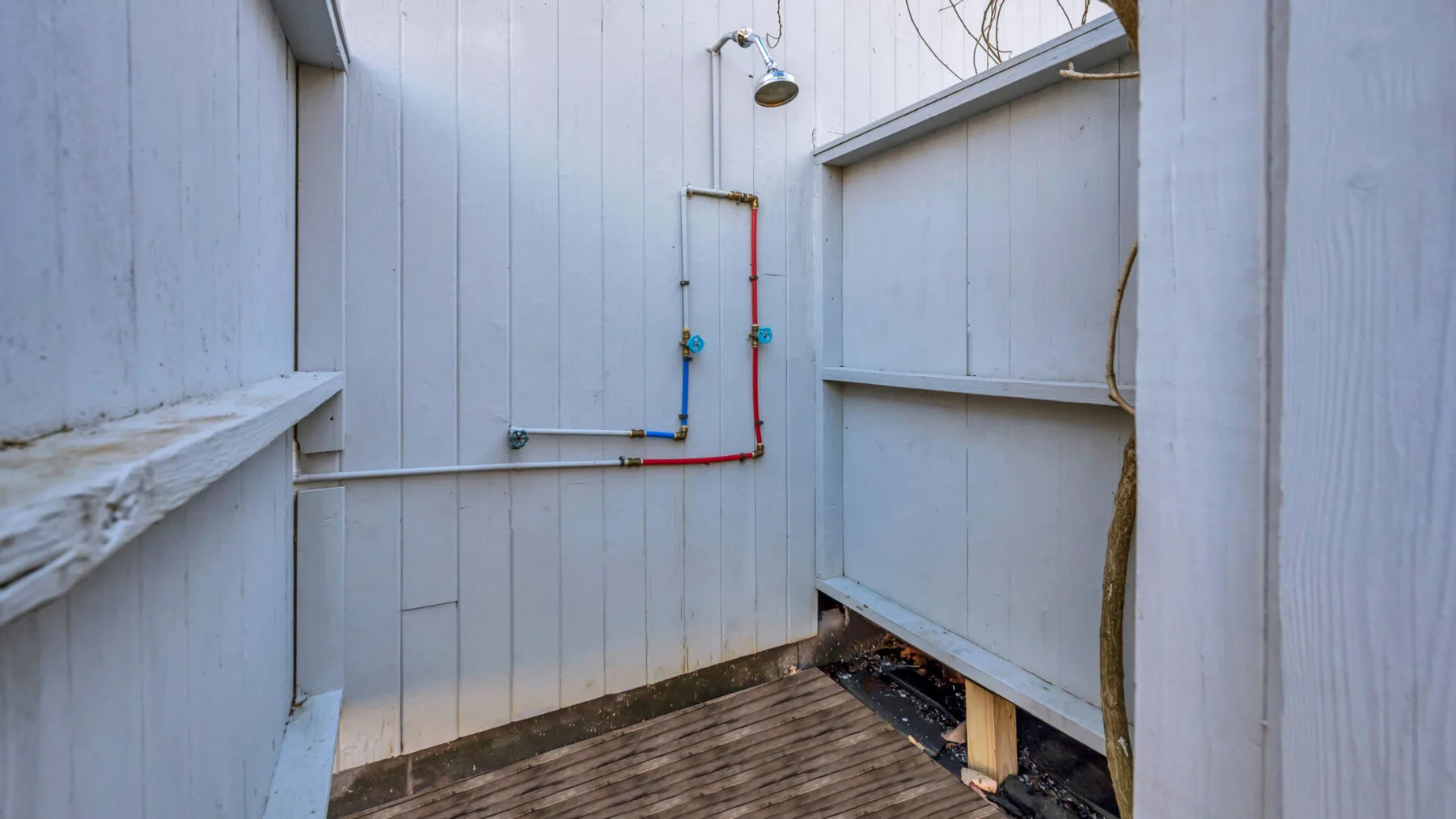 Una pequeña zona de ducha exterior con un cabezal de pared y tuberías expuestas.