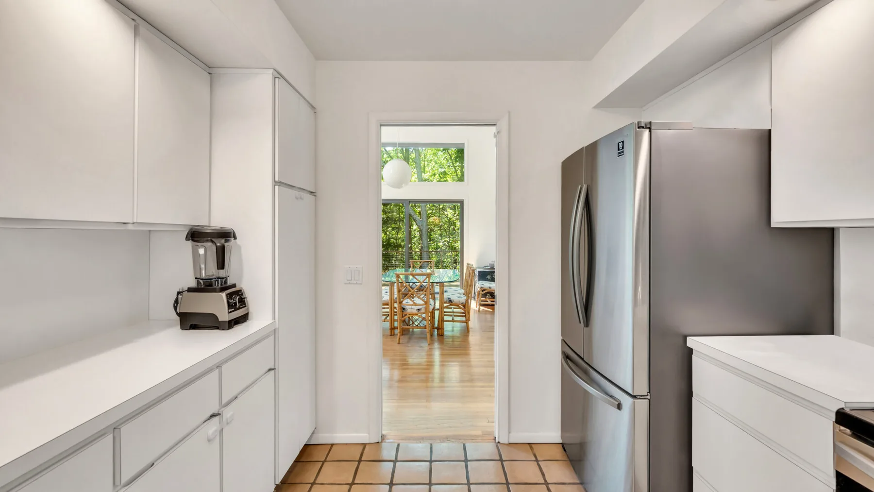 Una cocina luminosa con gabinetes blancos, un refrigerador y un comedor visible a través de la puerta.