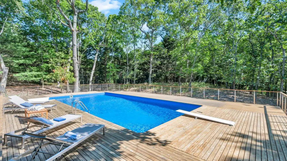 Una piscina exterior está junto a una terraza de madera rodeada de árboles.