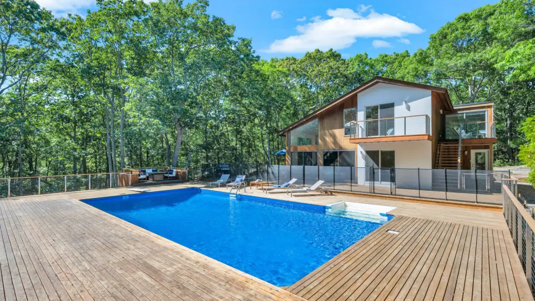 Una piscina al aire libre y una terraza de madera están junto a una casa moderna rodeada de árboles.