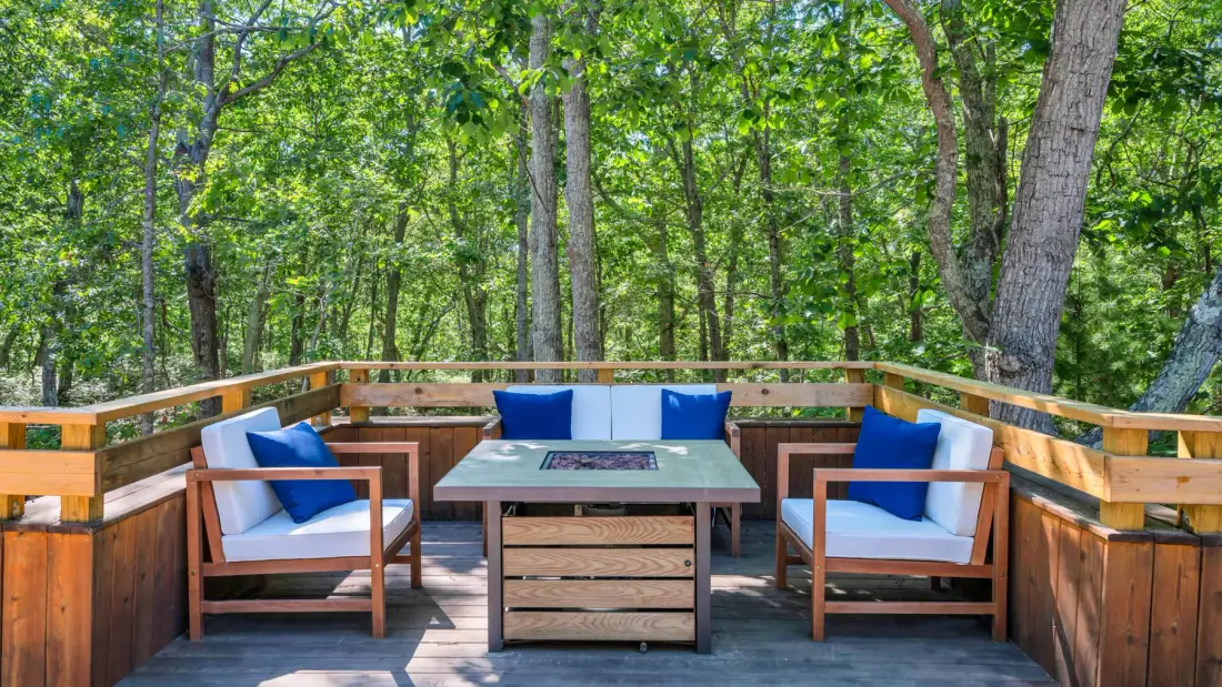 Una terraza de madera con asientos y una mesa al aire libre está rodeada de árboles frondosos.