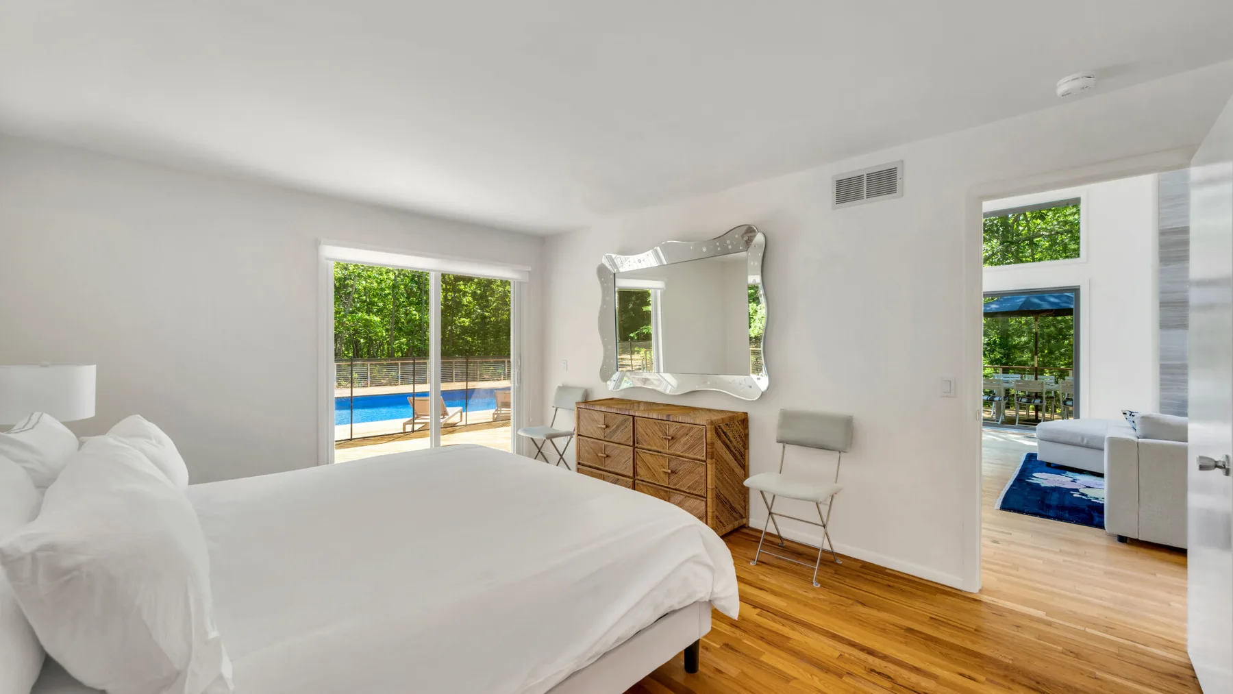 Un dormitorio luminoso con una cama grande, una cómoda de madera y ventanas con vistas a una piscina.