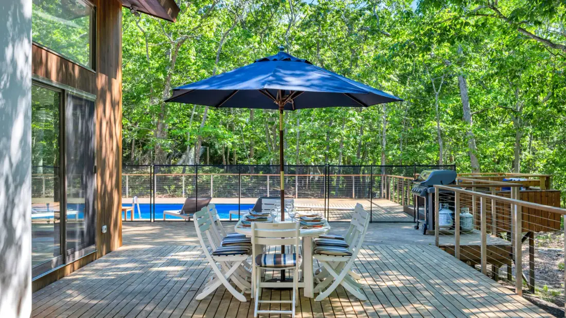Una terraza de madera tiene asientos al aire libre bajo una sombrilla azul, junto a una piscina cercada.