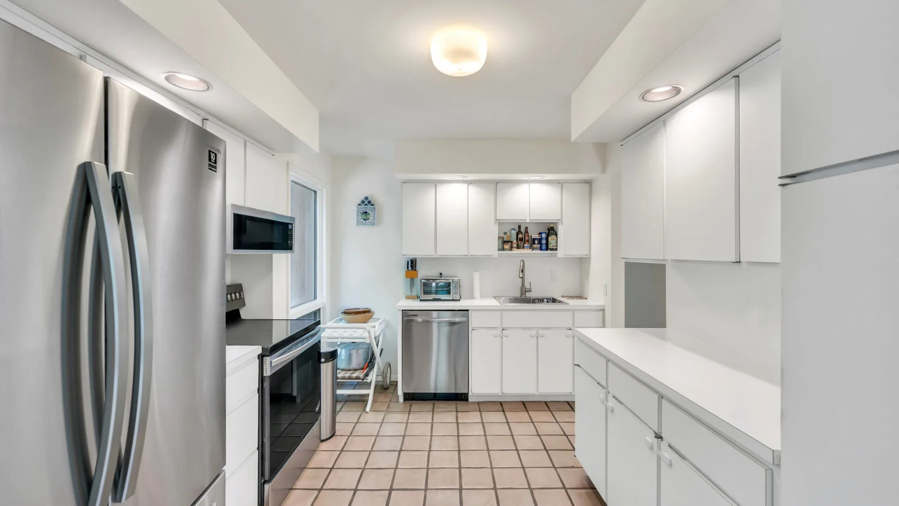 Una cocina luminosa con gabinetes blancos, electrodomésticos de acero inoxidable y piso de baldosas.
