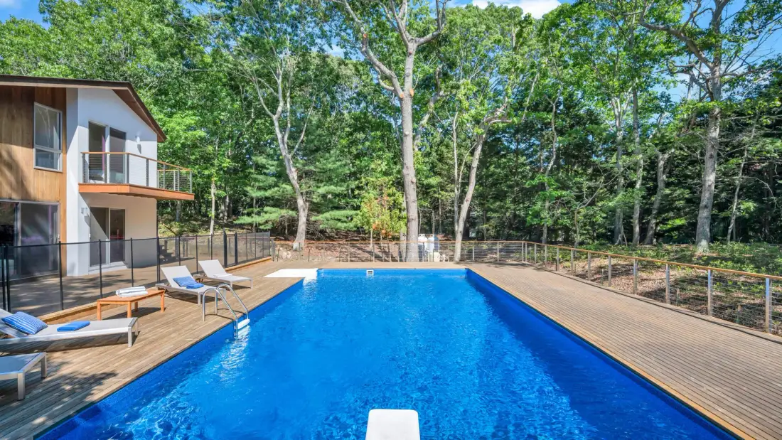 Una piscina rectangular está junto a una terraza de madera y una casa moderna.