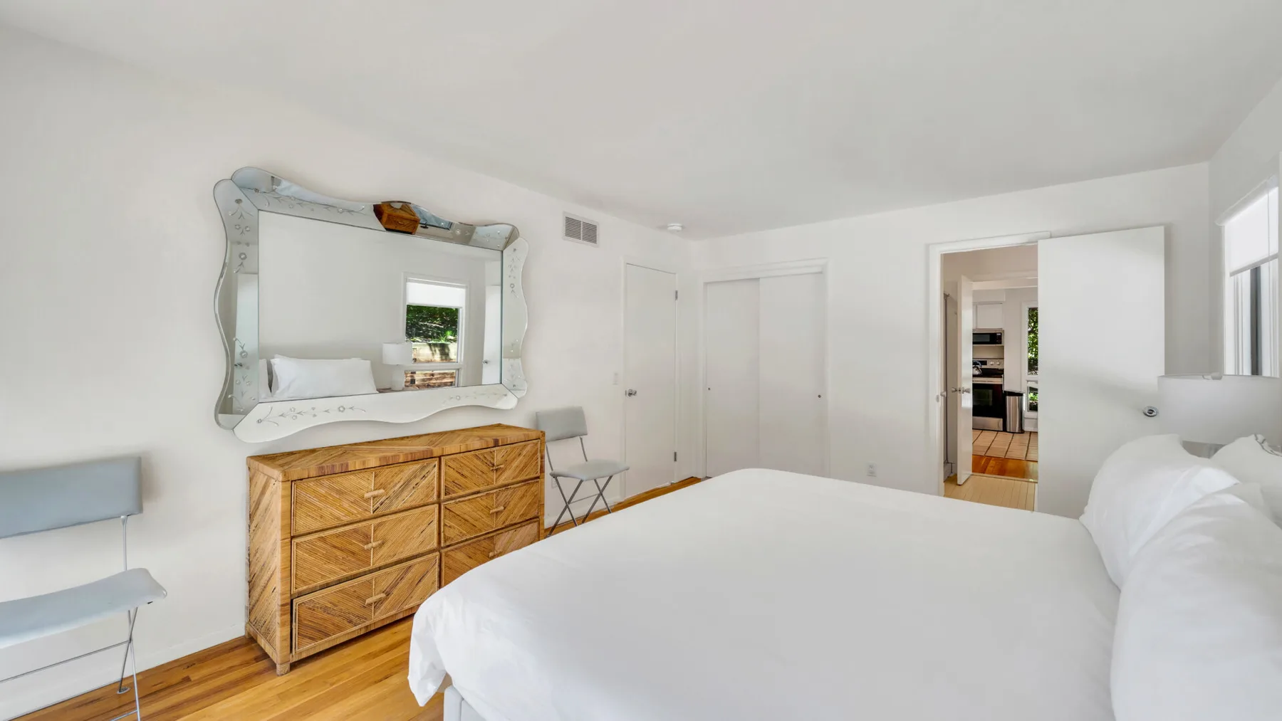 Un dormitorio luminoso con una cama grande, una cómoda de madera y un espejo de pared.