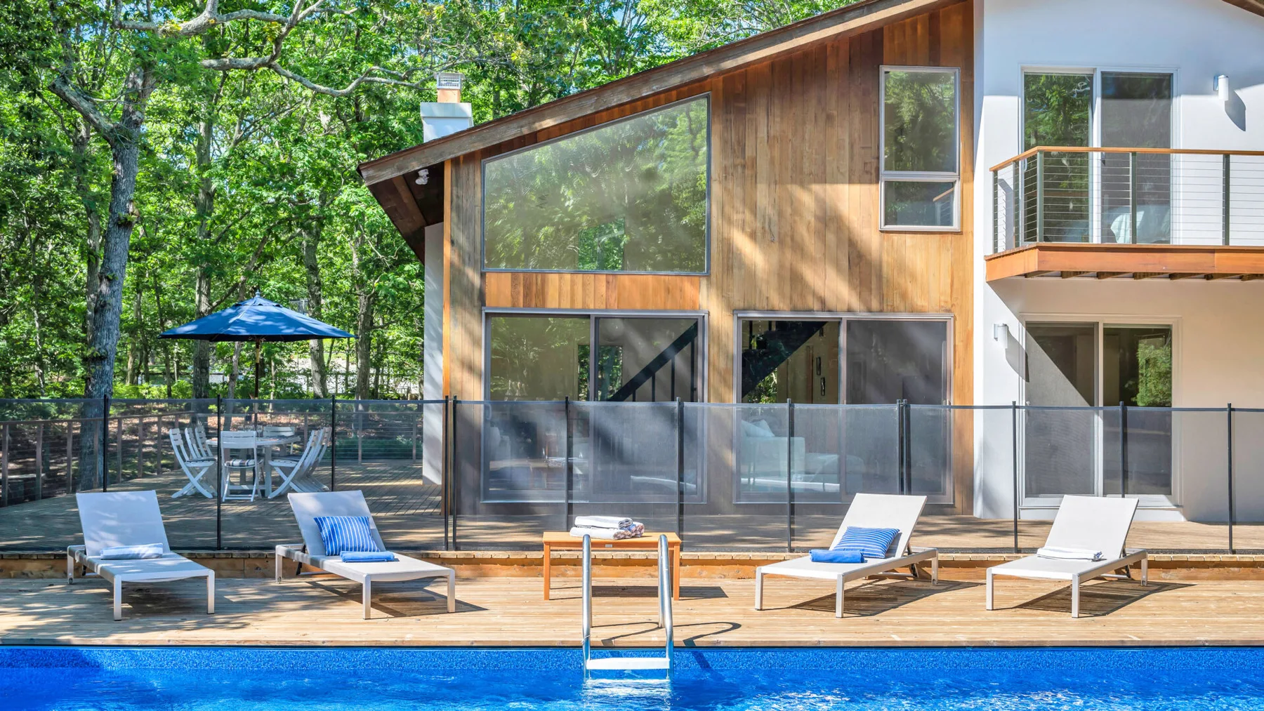 Una casa moderna da a una terraza junto a la piscina, con asientos y árboles frondosos cerca.