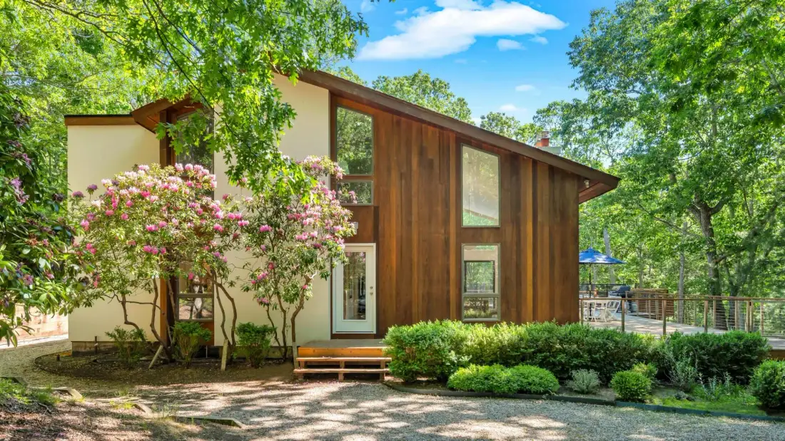 Exterior de una casa moderna con ventanas altas, árboles y arbustos en flor.