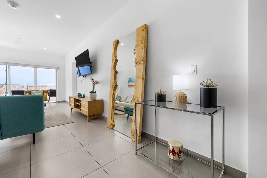 Step into your bright, open living space where natural wood accents and sleek modern furnishings create a welcoming coastal retreat. Through the glass doors, you'll glimpse the ocean beyond—your perfect escape.