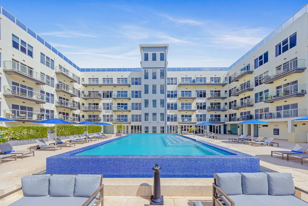 A sun-soaked courtyard pool stretches before modern terraced balconies, offering a tranquil retreat with ample lounge seating and shaded umbrellas beneath clear blue skies.